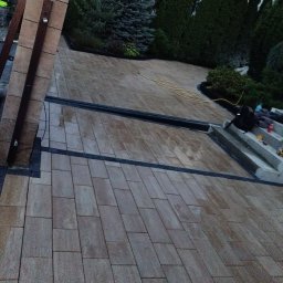 Roman Luhovyj - Ułożona kostka brukowa w odcieniach beżu i szarości, z czarnymi akcentami oddzielającymi strefy. Widoczne schody i elementy dekoracyjne z roślinnością. W tle iglaki.