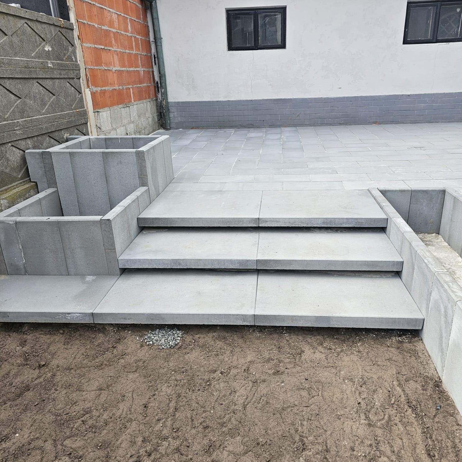 Nowoczesne schody z szarej kostki brukowej prowadzące na taras. Widoczne donice z tego samego materiału i fragment ogrodu z ziemią. Minimalistyczny design.