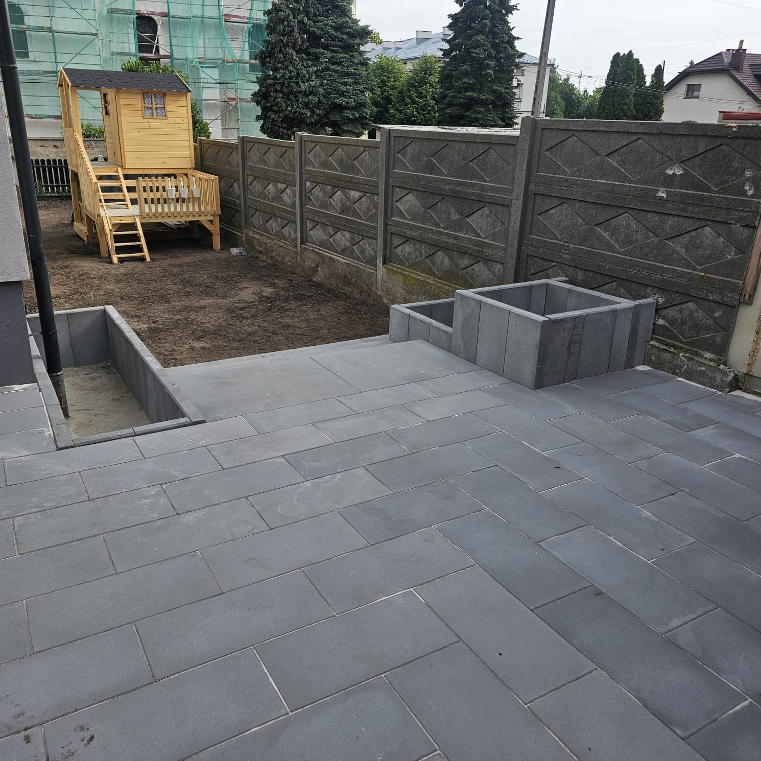 Szara, prostokątna kostka na tarasie z donicą i schodami, w tle drewniany domek dla dzieci i betonowe ogrodzenie. Ukończona realizacja.