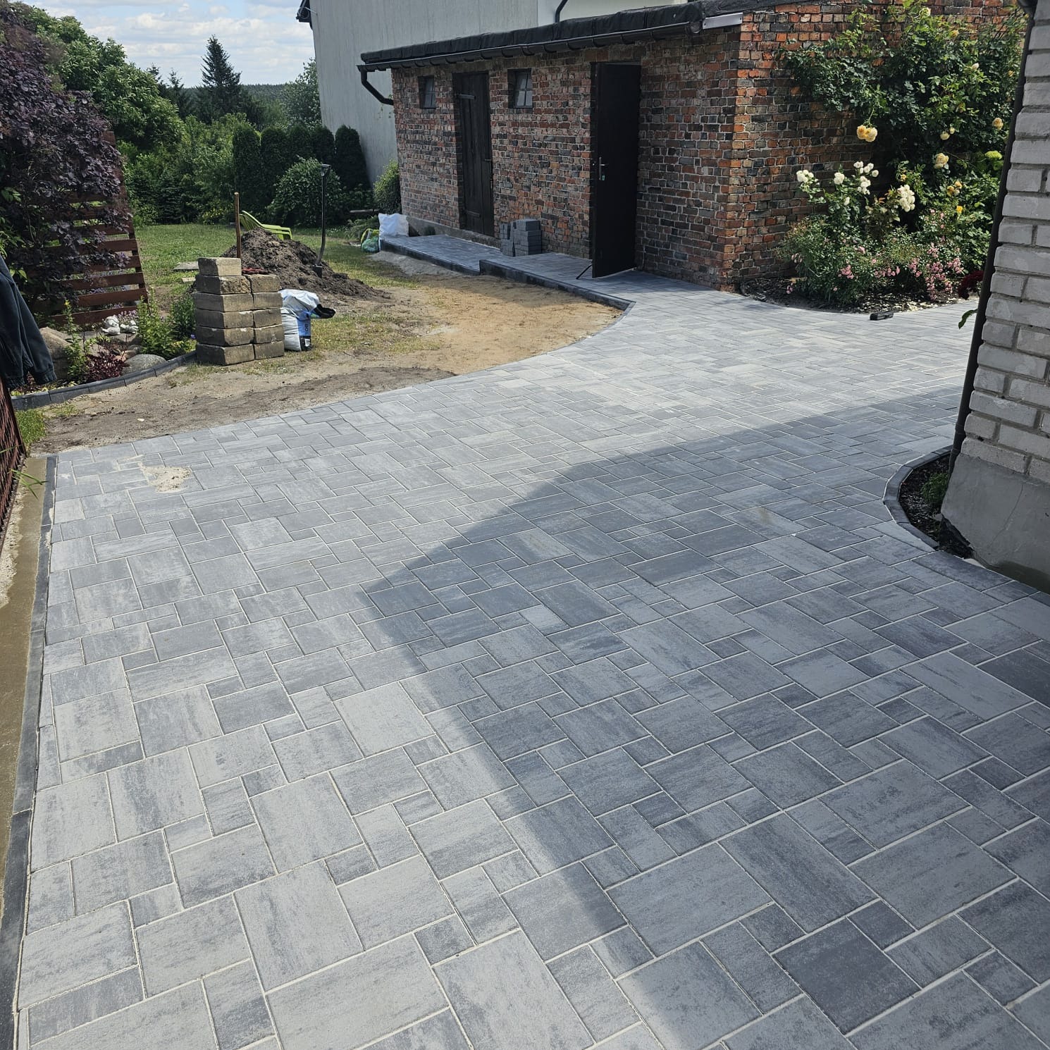 Szara kostka brukowa ułożona na podjeździe, z widocznym budynkiem gospodarczym z cegły i przygotowanym terenem pod dalsze prace brukarskie.