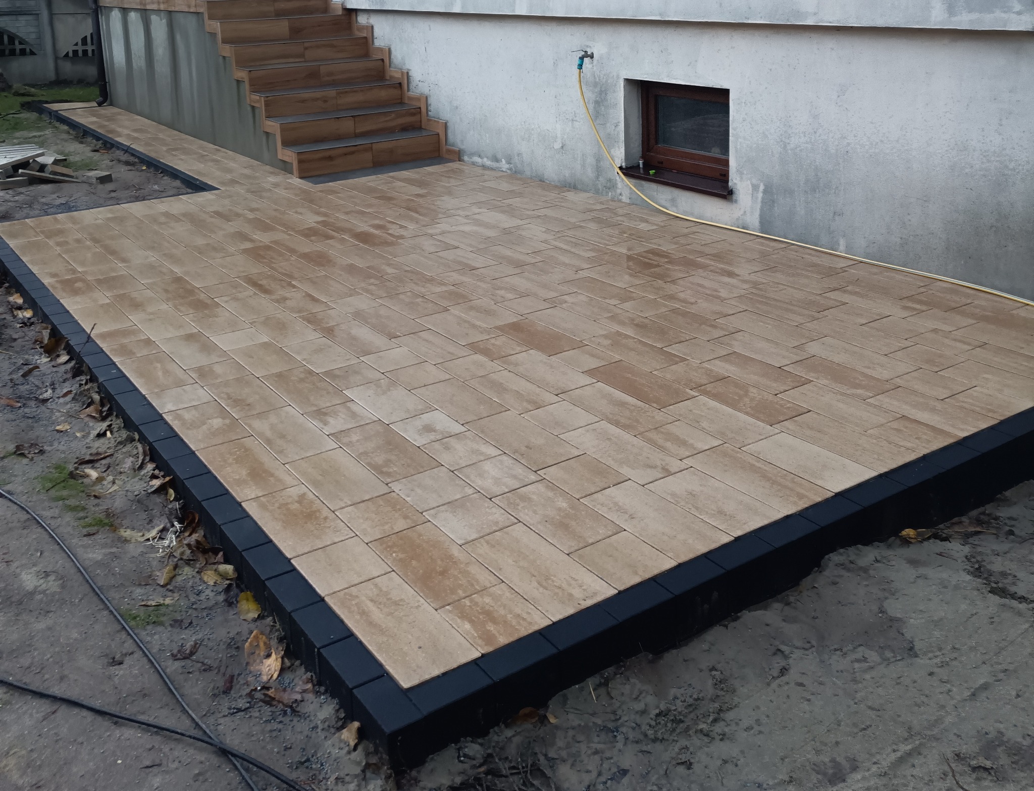 Ułożona, beżowa kostka brukowa z czarnym obrzeżem tworzy taras przy budynku z drewnianymi schodami. Widoczny kran i okno piwniczne.