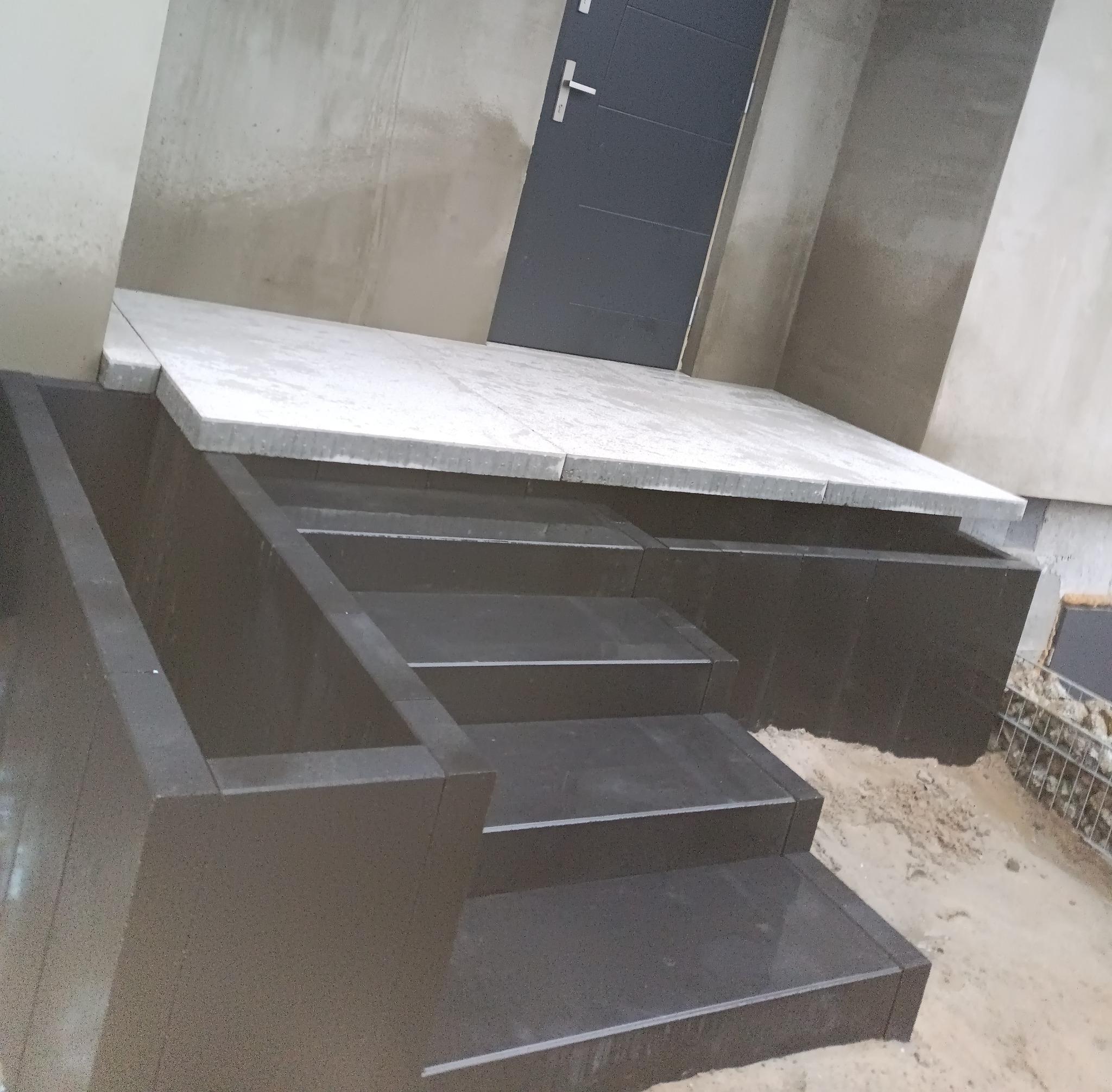 Granitowe schody wejściowe w trakcie montażu, z widocznym podestem i surowym otoczeniem. Detal układania kostki granitowej, ciemny kamień, jasny podest.