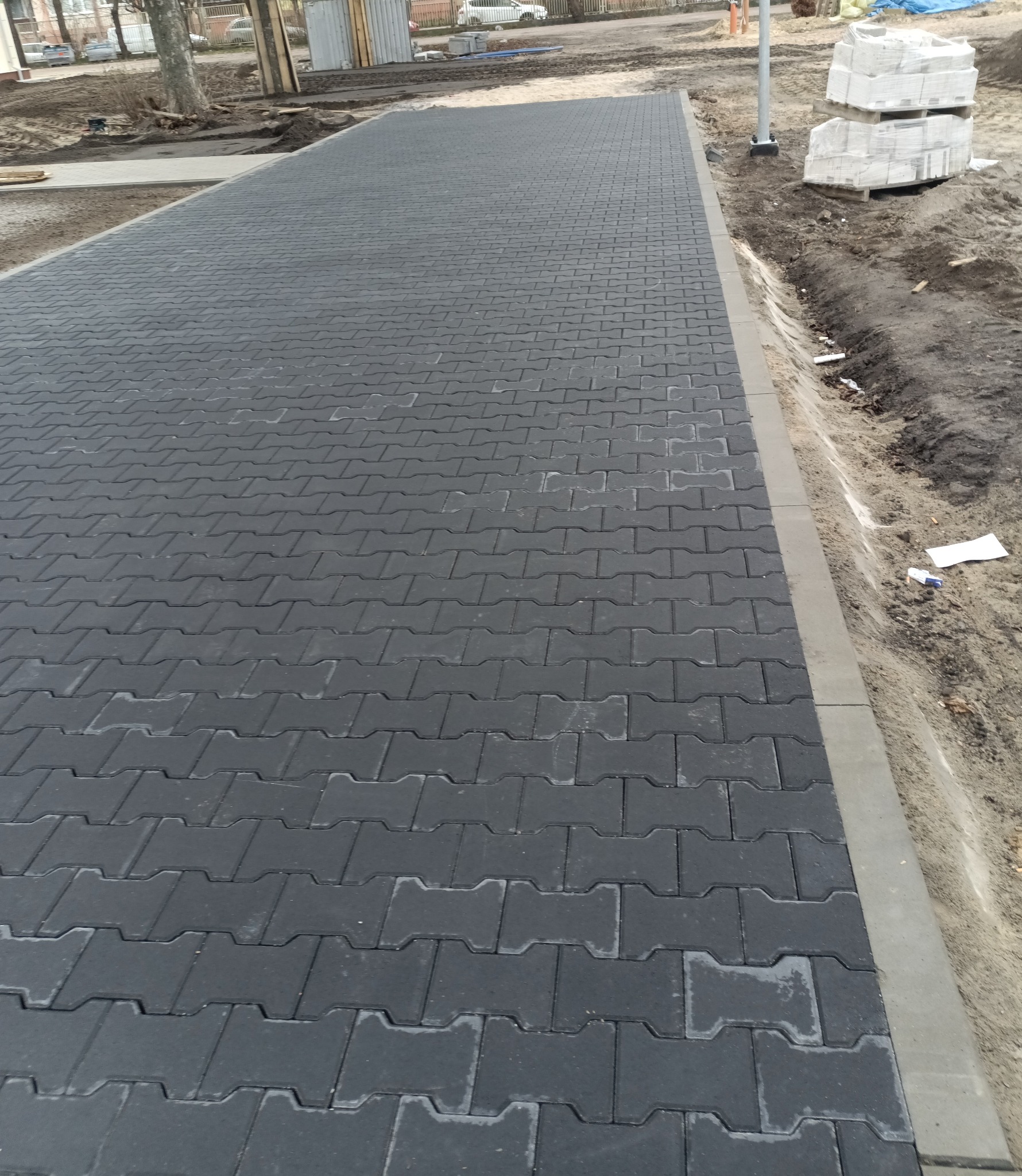 Świeżo ułożona, szara kostka brukowa typu 'I' tworząca chodnik. W tle widoczne palety z kostką i przygotowany teren.