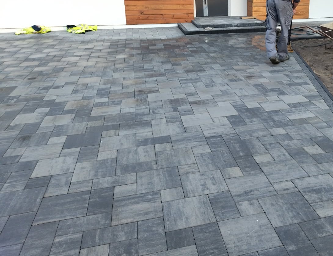 Szara kostka brukowa ułożona na podjeździe, widoczne ubrania robocze i fragment osoby. Nowoczesny wzór kostki, świeżo po ułożeniu, zarys domu w tle.