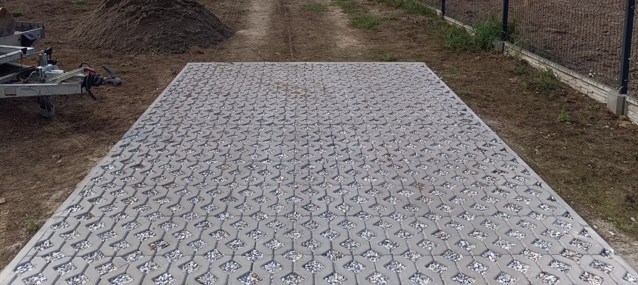 Ułożona szara ażurowa kostka brukowa wypełniona drobnym kamieniem na podwórku, widok z góry, w tle przyczepka i hałda ziemi.