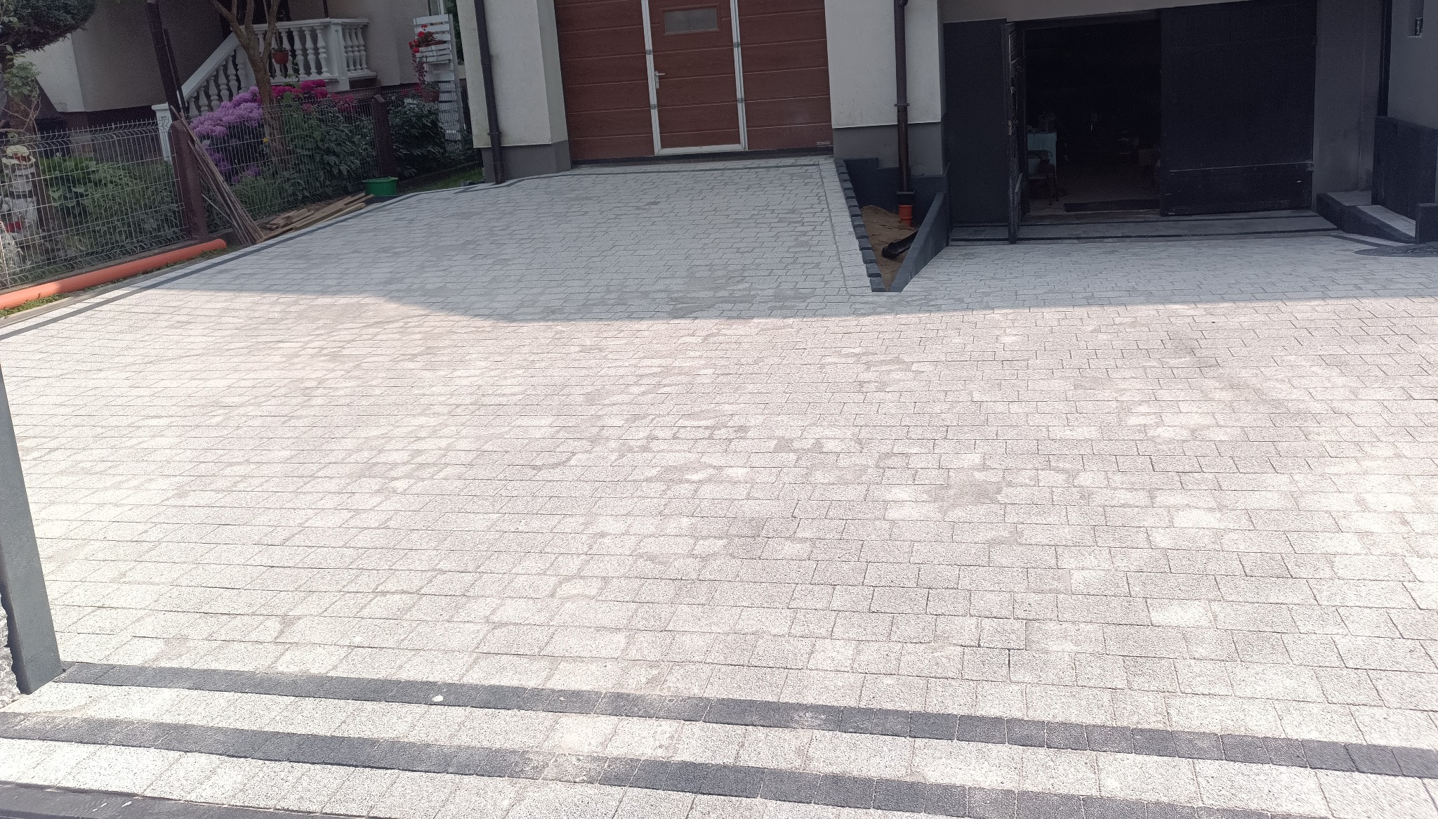 Jasnoszara kostka brukowa ułożona na podjeździe, z ciemnoszarymi obrzeżami i schodami. W tle budynek z garażem i roślinność.