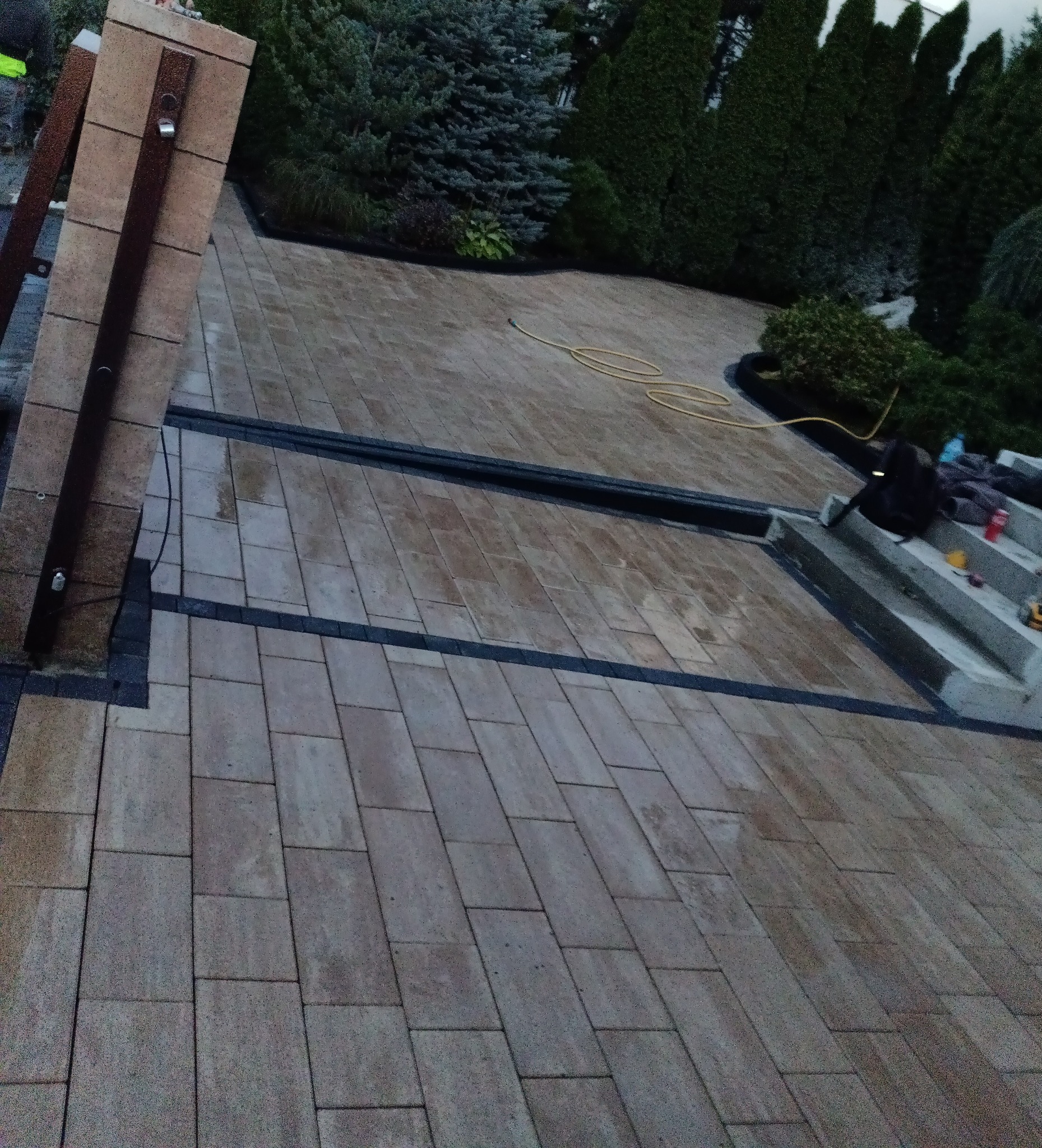 Ułożona kostka brukowa w odcieniach beżu i szarości, z czarnymi akcentami oddzielającymi strefy. Widoczne schody i elementy dekoracyjne z roślinnością. W tle iglaki.