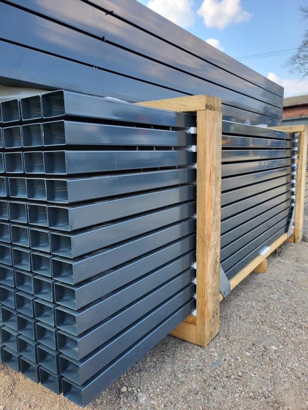 Stos szarych, metalowych profili kwadratowych na drewnianej palecie, prawdopodobnie elementy konstrukcyjne ogrodzenia, widoczne niebo i budynek w tle.