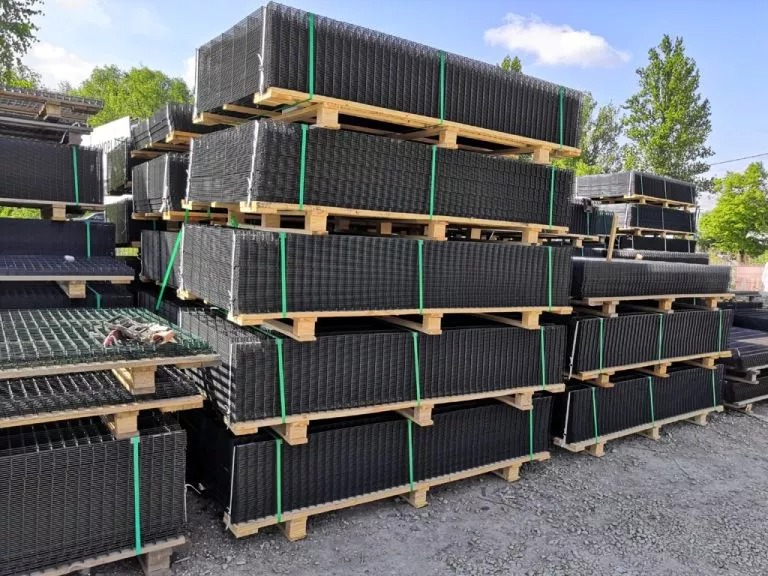 Stos palet z panelami ogrodzeniowymi w czarnym kolorze, zabezpieczonych zielonymi taśmami, na tle zieleni i błękitnego nieba. Widok z dołu.