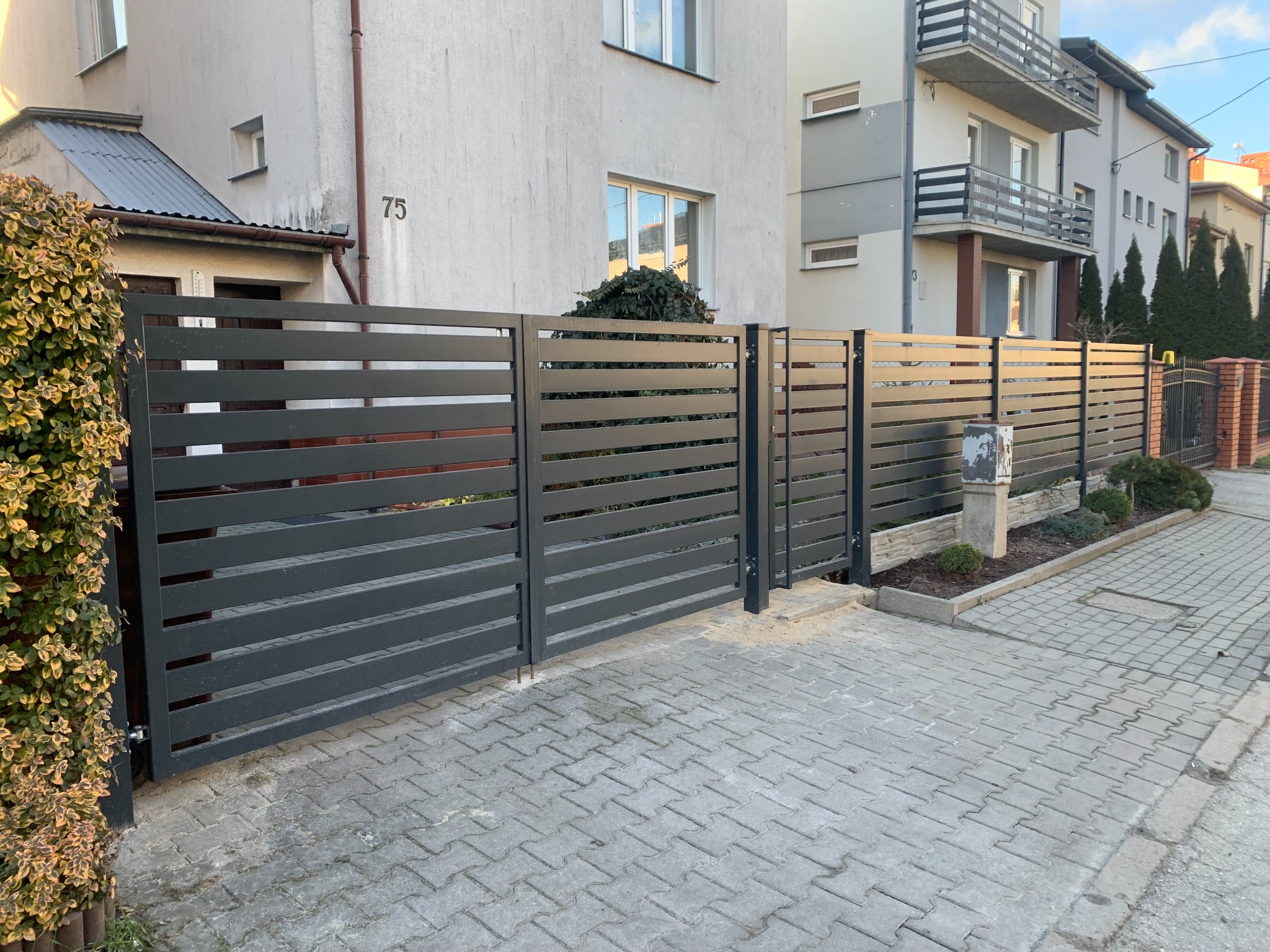 Nowoczesne, poziome przęsła ogrodzeniowe w kolorze grafitowym, oddzielające posesję od chodnika z kostki brukowej. Widoczny fragment budynku mieszkalnego w tle.