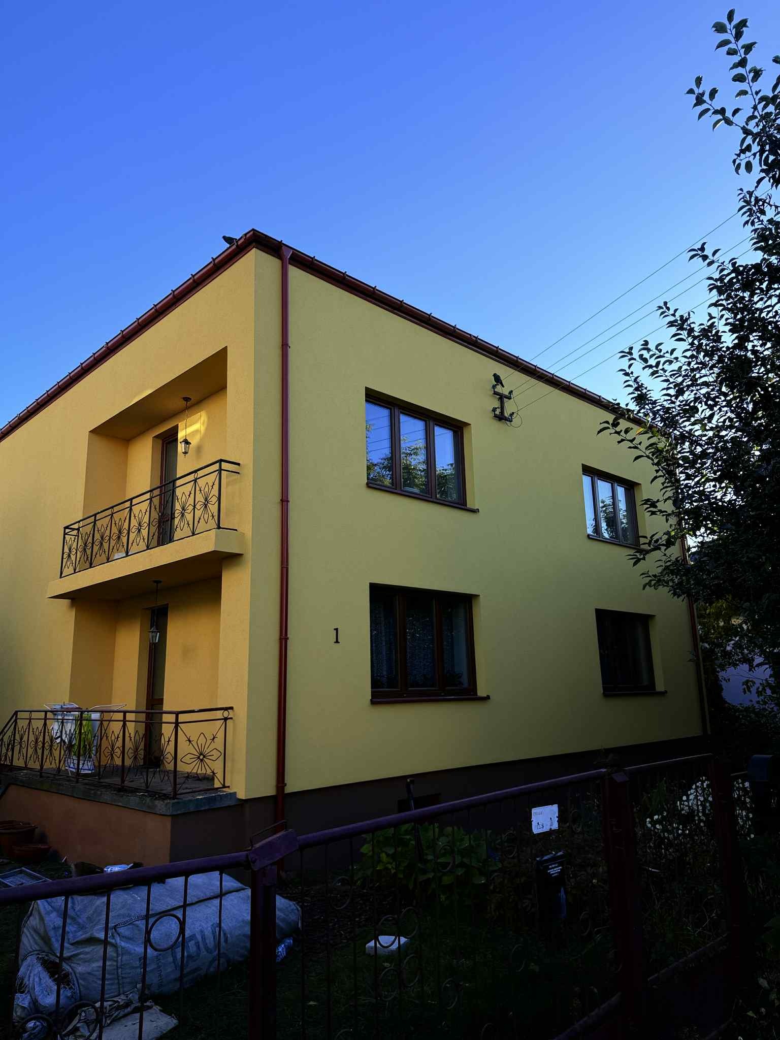Żółty, dwupiętrowy dom z balkonem i brązowymi oknami, widziany z dołu pod kątem. Na elewacji widoczny numer '1'. Ciemne tło z zielenią i niebieskim niebem.