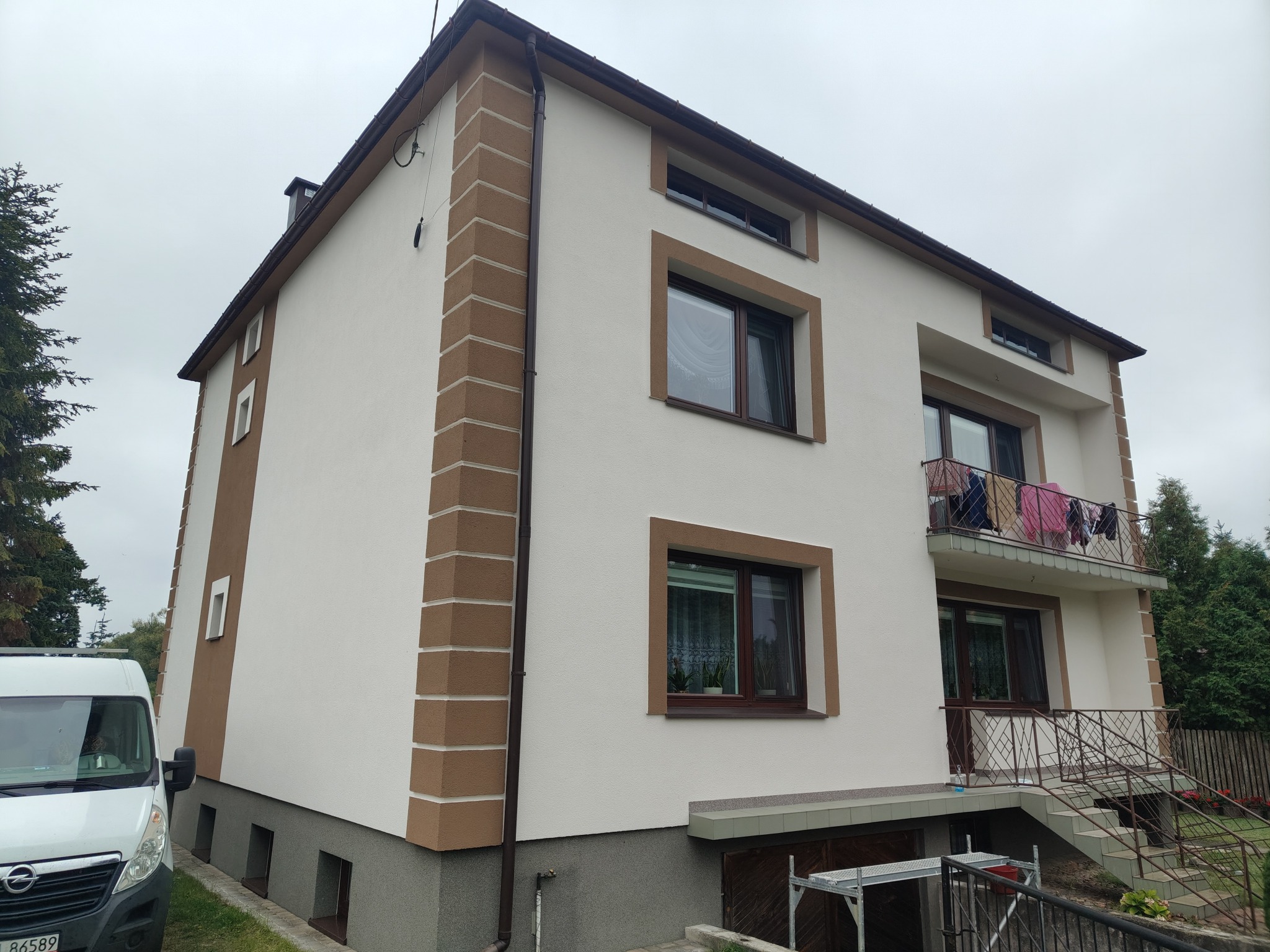 Dwukondygnacyjny dom z elewacją w odcieniach bieli i brązu, z widocznym balkonem i oknami z brązowymi ramami. Na pierwszym planie zaparkowany biały bus.