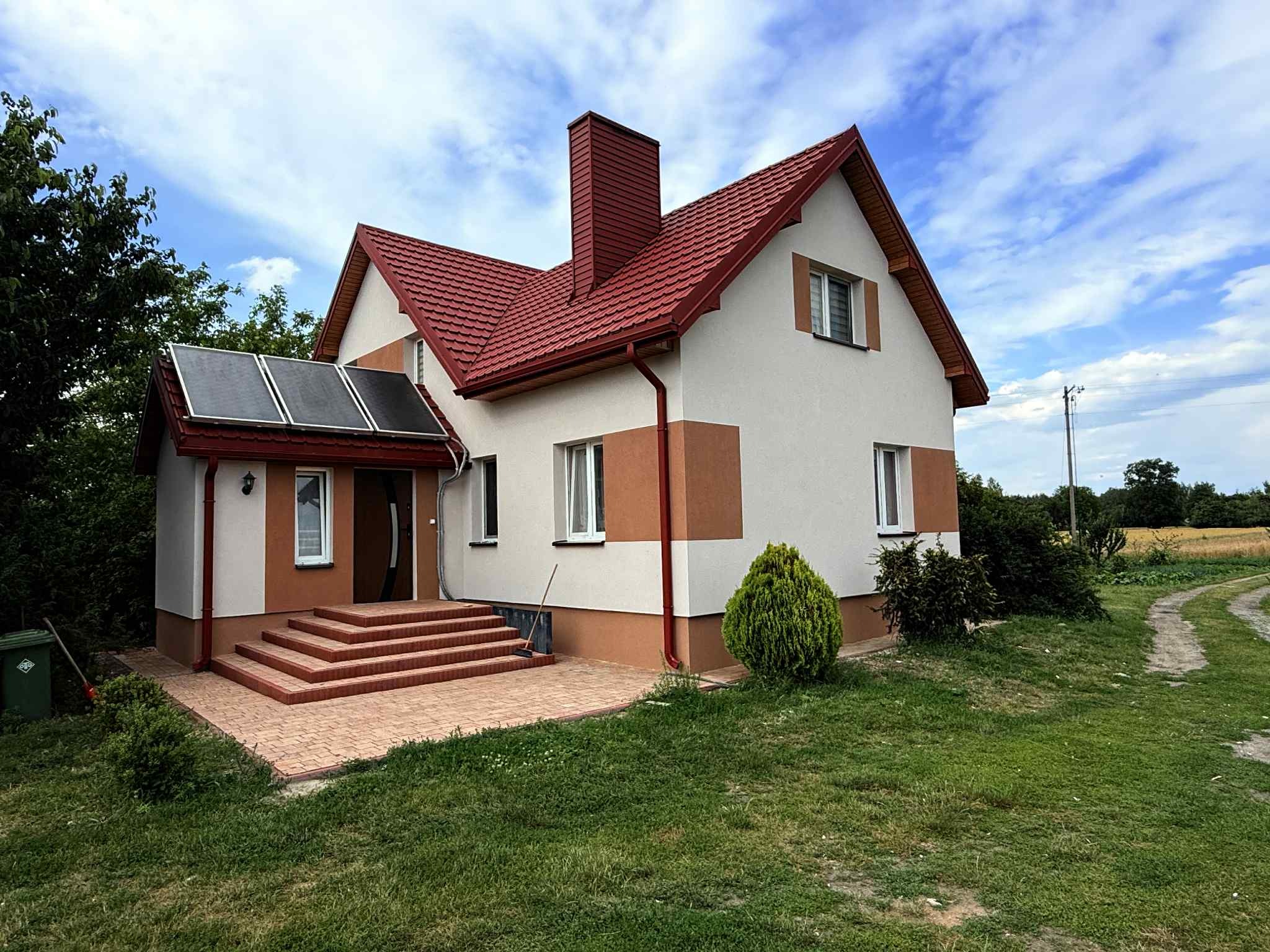 Dwukondygnacyjny dom z czerwonym dachem i panelami słonecznymi, otoczony zielonym trawnikiem i ścieżką. Jasna elewacja z brązowymi akcentami. Widok z poziomu gruntu.