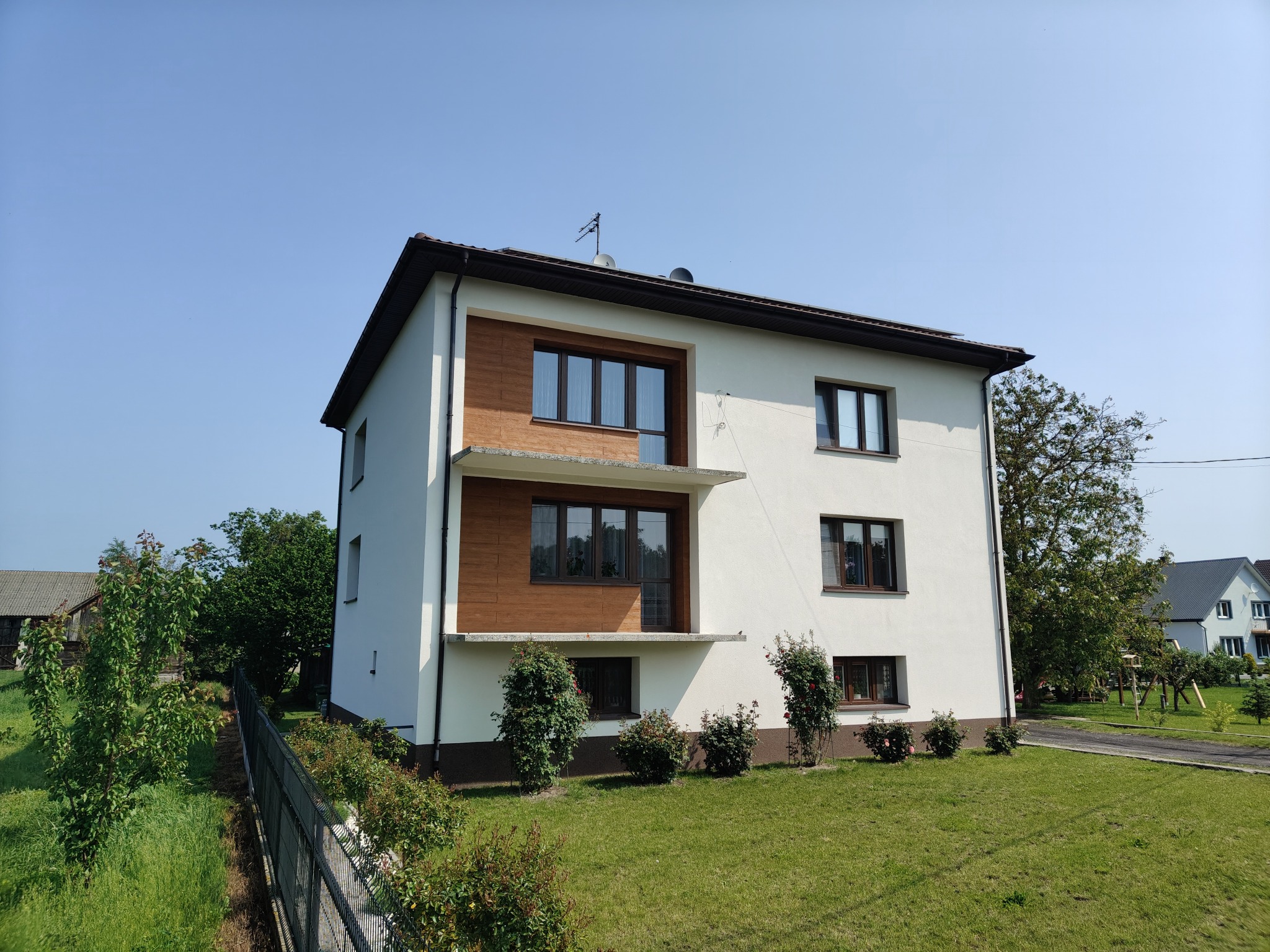 Dwupiętrowy dom z białą elewacją i drewnianymi akcentami, widoczny z perspektywy ogrodu. Nowoczesny design z balkonami i zadbanym trawnikiem.