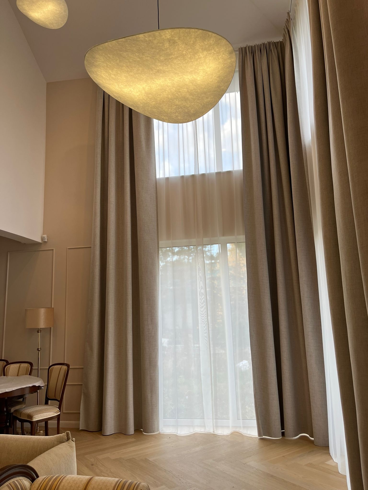 Wysokie okno z beżowymi zasłonami i delikatną firaną w przestronnym salonie z parkietem w jodełkę. Nad oknem designerska lampa w kształcie chmury.