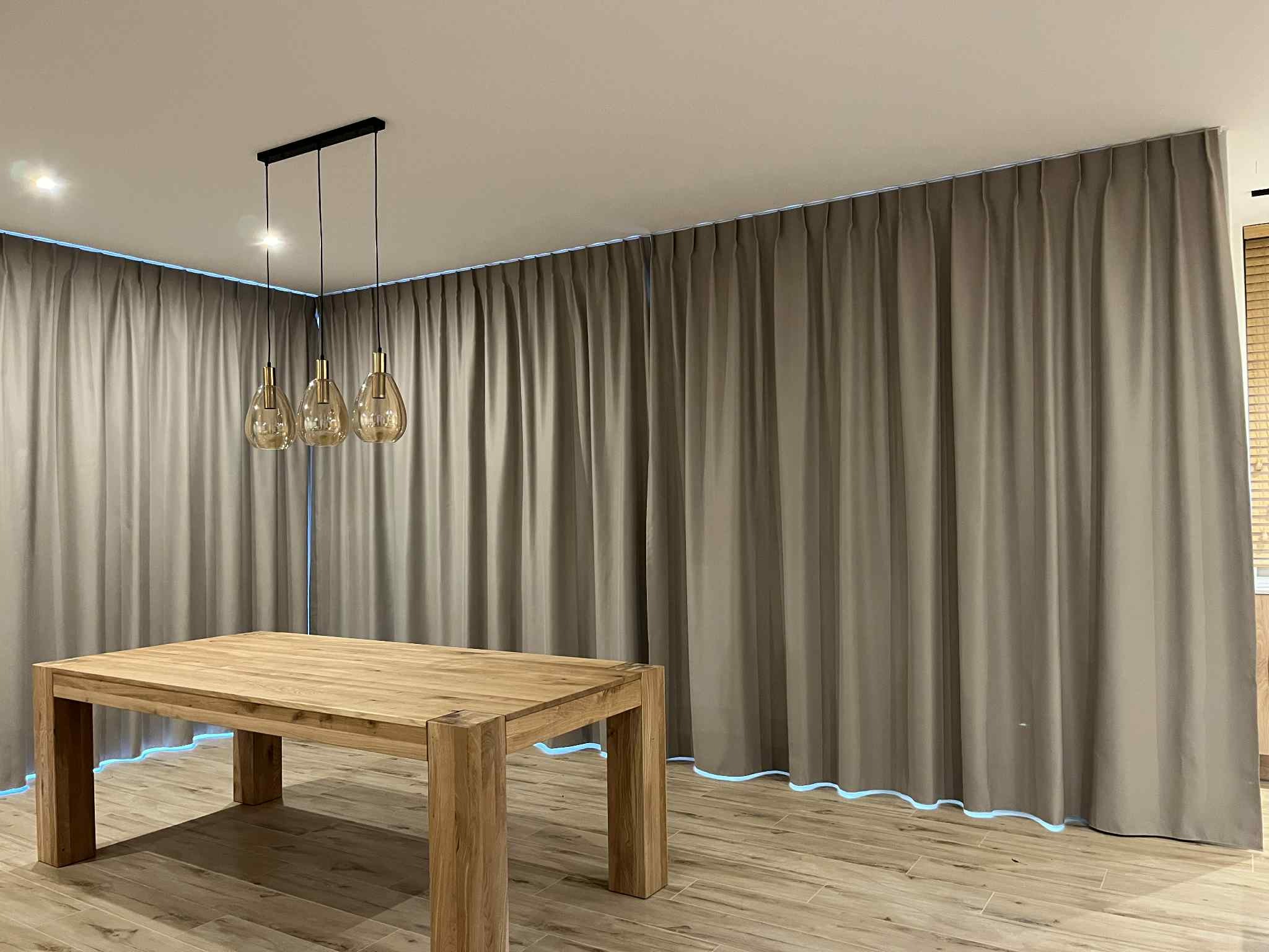 Beżowe zasłony z podświetleniem LED wzdłuż krawędzi, wiszące od sufitu do podłogi, z drewnianym stołem na pierwszym planie i trzema lampami wiszącymi nad nim.
