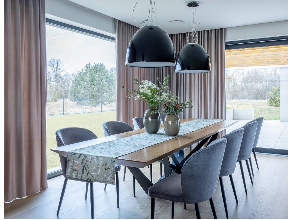 Elegancka jadalnia z długim stołem, szarymi krzesłami i beżowymi zasłonami w tle okna z widokiem na zieleń. Dwie czarne lampy wiszą nad stołem.