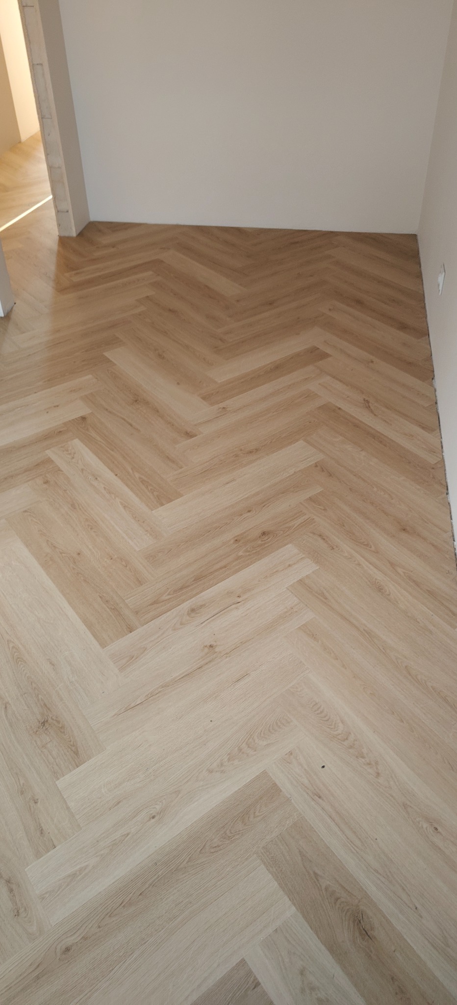 Jasne panele ułożone w jodełkę francuską, widok z góry. Minimalistyczne wnętrze z białymi ścianami i otwartym przejściem do innego pomieszczenia.