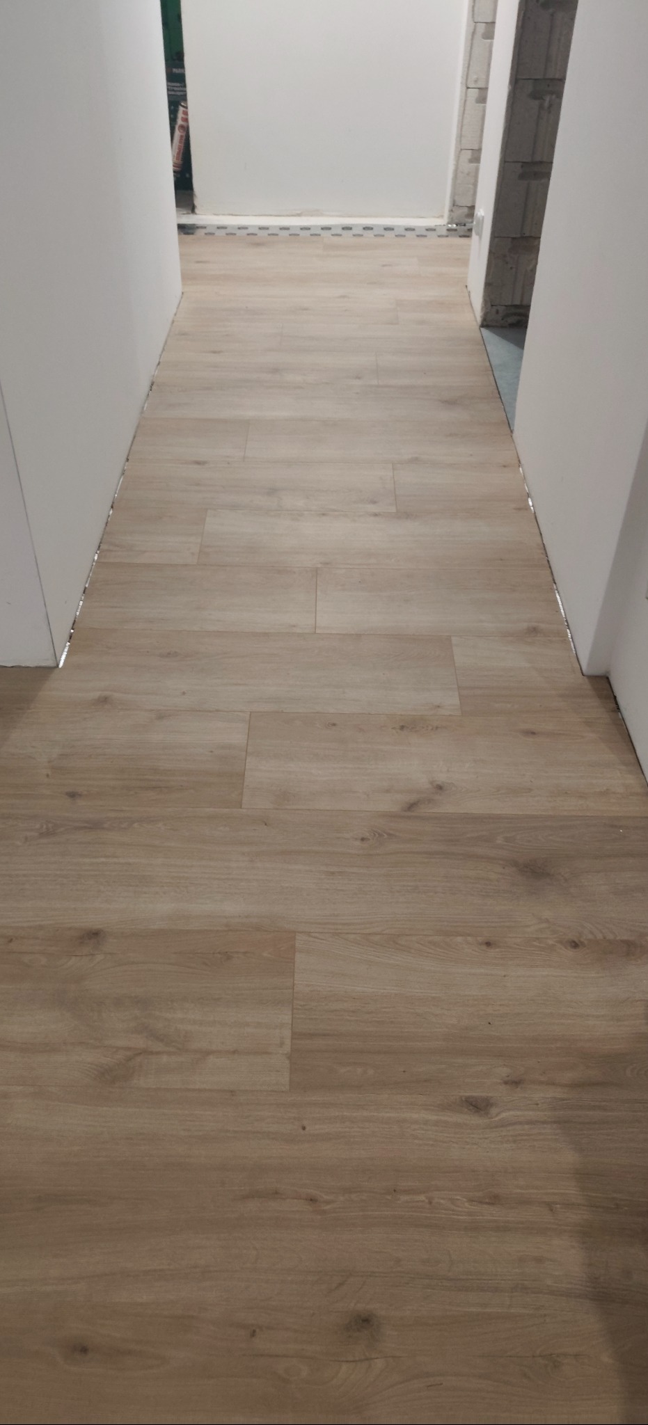 Jasne panele podłogowe ułożone w korytarzu, widok z perspektywy wejścia. Minimalistyczne białe ściany i brak listew przypodłogowych podkreślają nowoczesny charakter.