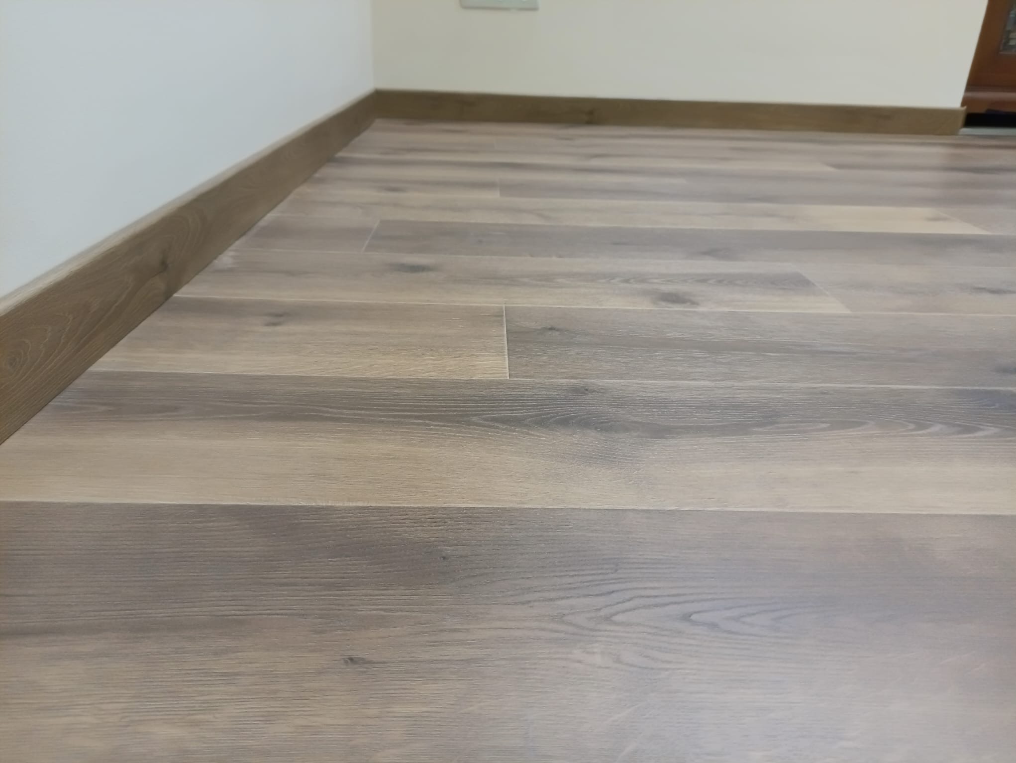 Ułożone panele podłogowe w odcieniach szarości i brązu, z widoczną listwą przypodłogową w kolorze drewna. Neutralne tło ściany i fragment mebla w tle.