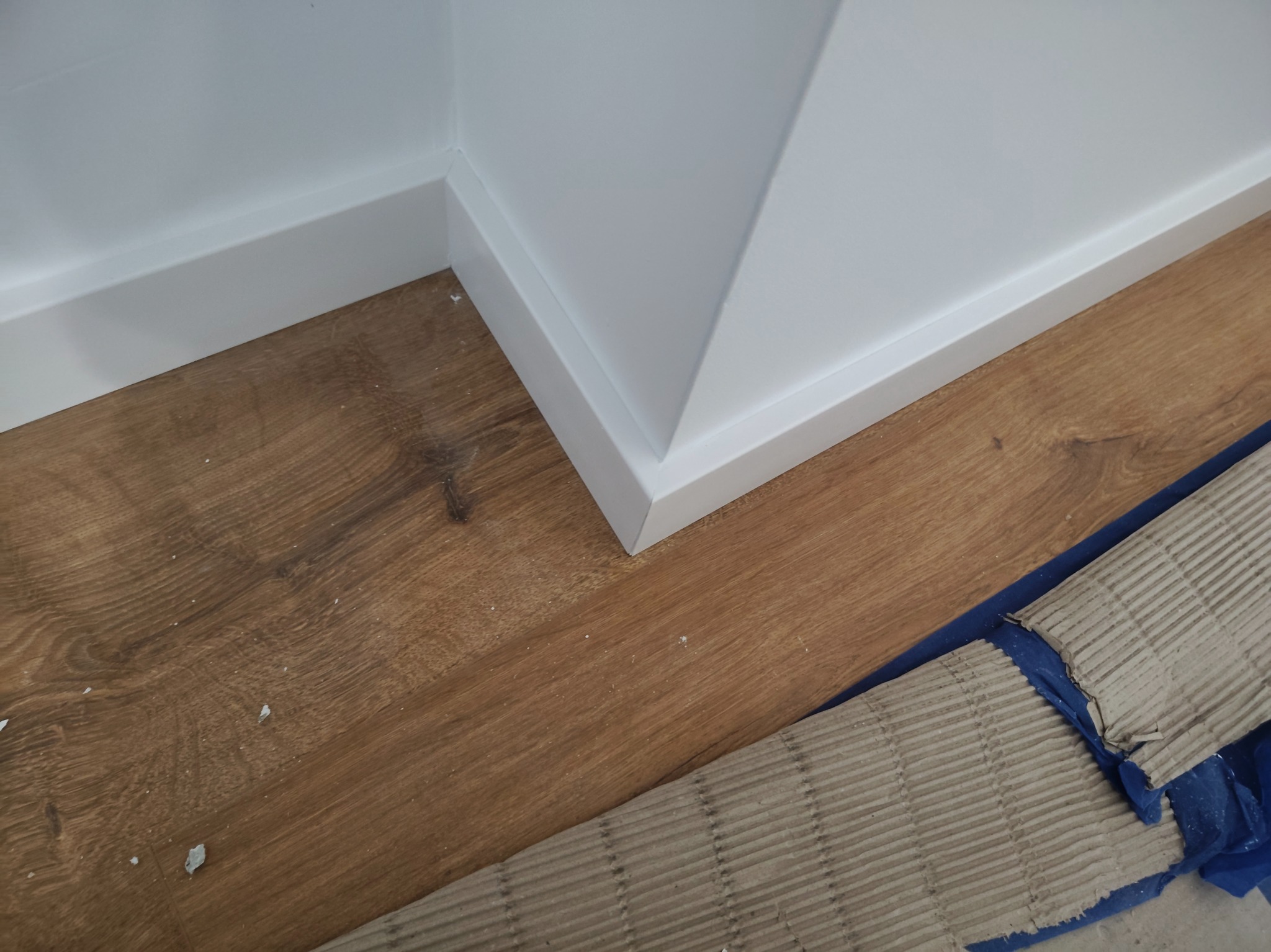 Detal wykończenia: brązowe panele podłogowe z listwami przypodłogowymi w kolorze białym, zabezpieczone kartonem falistym i folią malarską przy ścianie.