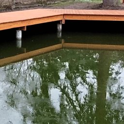 LUMI-ART Michał Oleksik - Drewniany taras nad stawem, wsparty na betonowych słupach. Widoczne odbicie konstrukcji w wodzie, otoczenie naturalne z drzewami i krzewami. Wykończenie w ciepłym odcieniu brązu.