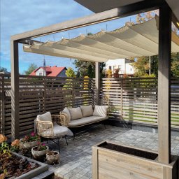 Nowoczesna pergola z beżową, zwijaną markizą, otoczona drewnianym ogrodzeniem i roślinami w donicach. Wygodny zestaw wypoczynkowy na szarym bruku.