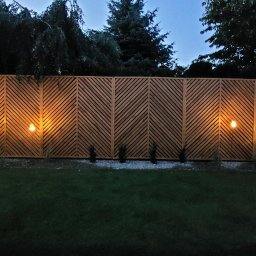LUMI-ART Michał Oleksik - Drewniane ogrodzenie z ukośnymi listwami, oświetlone lampami ogrodowymi, tworzące ciepłą atmosferę w ogrodzie o zmierzchu. Widoczne drzewa w tle.