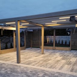 LUMI-ART Michał Oleksik - Nowoczesna pergola z szarymi słupami i oświetleniem LED, z roletami i dekoracyjnymi panelami bocznymi. Podłoże wyłożone jasnoszarą kostką brukową.