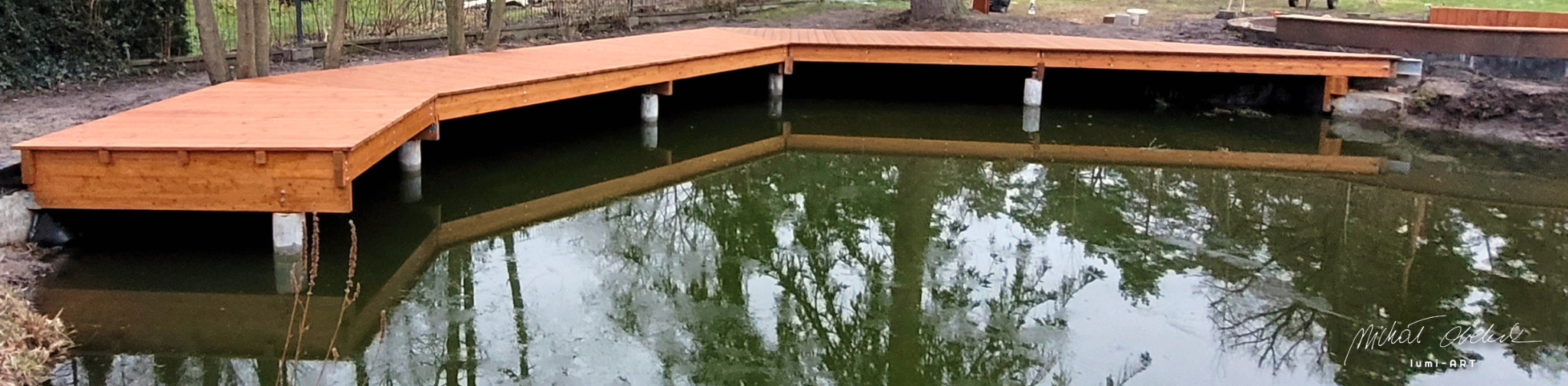 Drewniany taras nad stawem, wsparty na betonowych słupach. Widoczne odbicie konstrukcji w wodzie, otoczenie naturalne z drzewami i krzewami. Wykończenie w ciepłym odcieniu brązu.