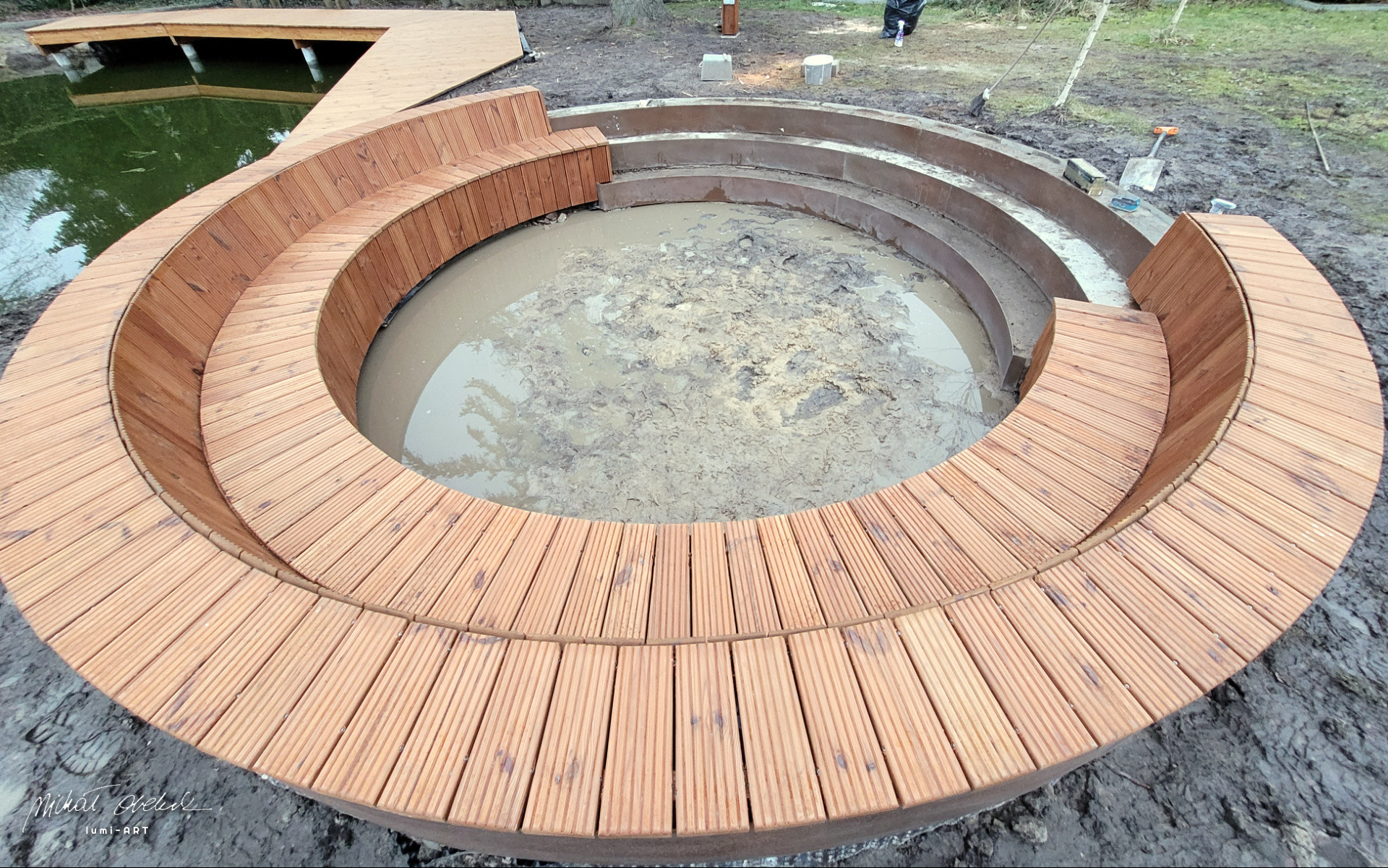 Okrągła, drewniana ławka otaczająca zagłębienie z wodą, obok drewniany pomost i błotniste podłoże. Nietypowy element architektury ogrodowej w trakcie budowy.