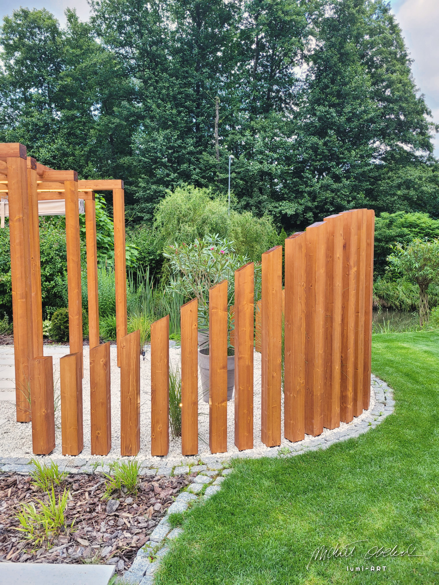 Nowoczesna pergola z drewna w ogrodzie, z elementami ażurowej konstrukcji i pionowymi deskami tworzącymi półokrągłą osłonę. Jasne drewno kontrastuje z zielenią trawy i drzew.