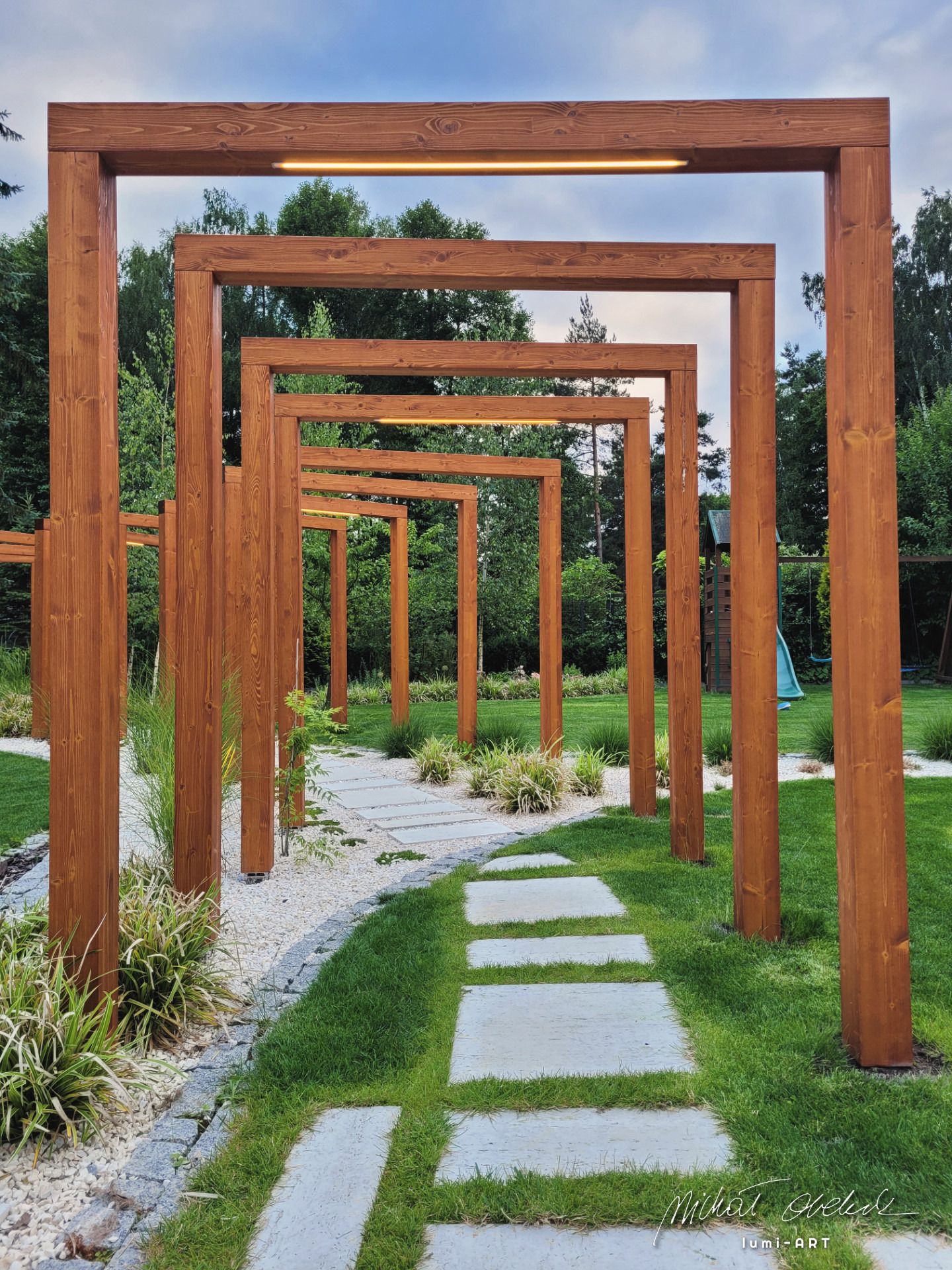 Drewniana pergola z oświetleniem LED, tworząca tunel nad ścieżką z płyt i białego żwiru w ogrodzie. Perspektywa z niskiego kąta, widoczne drzewa w tle.