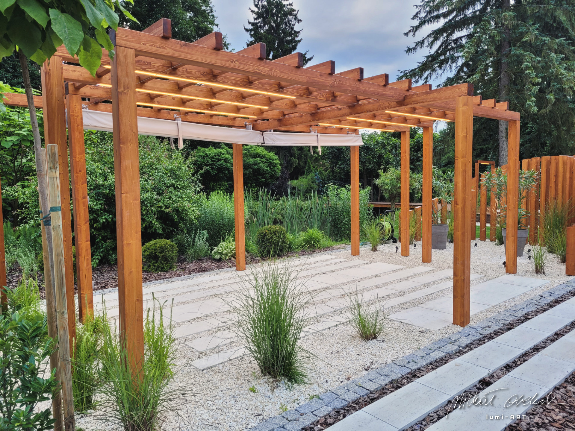 Drewniana pergola z oświetleniem LED i zwijanymi roletami, otoczona zielenią i białym żwirem, tworząca zacienioną strefę relaksu w ogrodzie. Widoczne elementy dekoracyjne i staranne wykończenie.