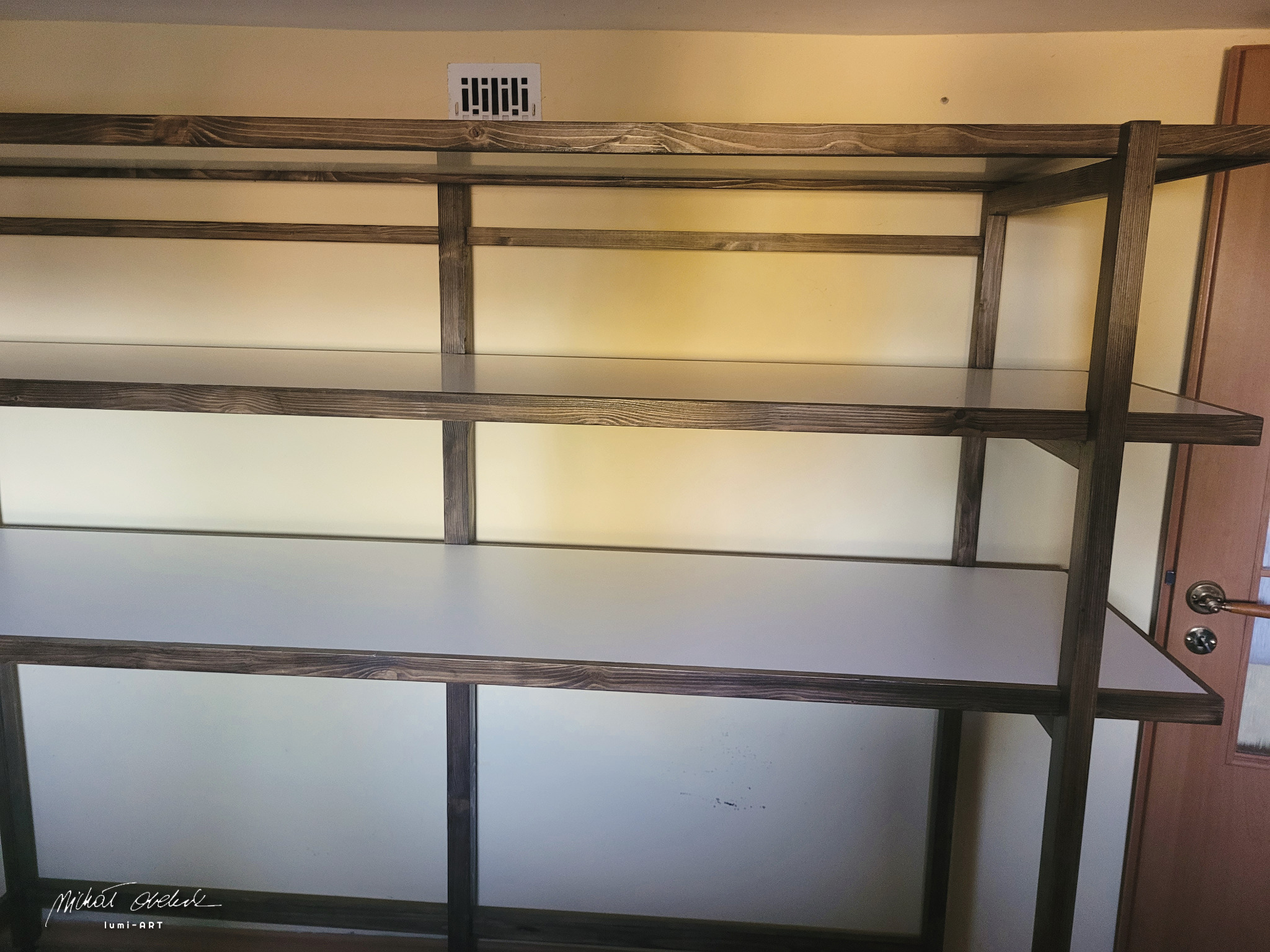 Ręcznie robiony regał z ciemnego drewna i białymi półkami, ustawiony przy ścianie z kratką wentylacyjną i drzwiami. Widoczne słoje drewna i detale konstrukcyjne.