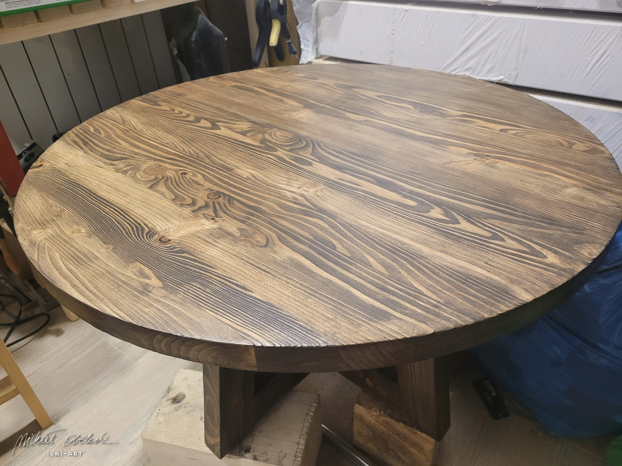 Okrągły, drewniany stół z wyraźnym usłojeniem, wykończony w ciemnym odcieniu brązu, na masywnej, drewnianej podstawie. Widoczne detale obróbki drewna.