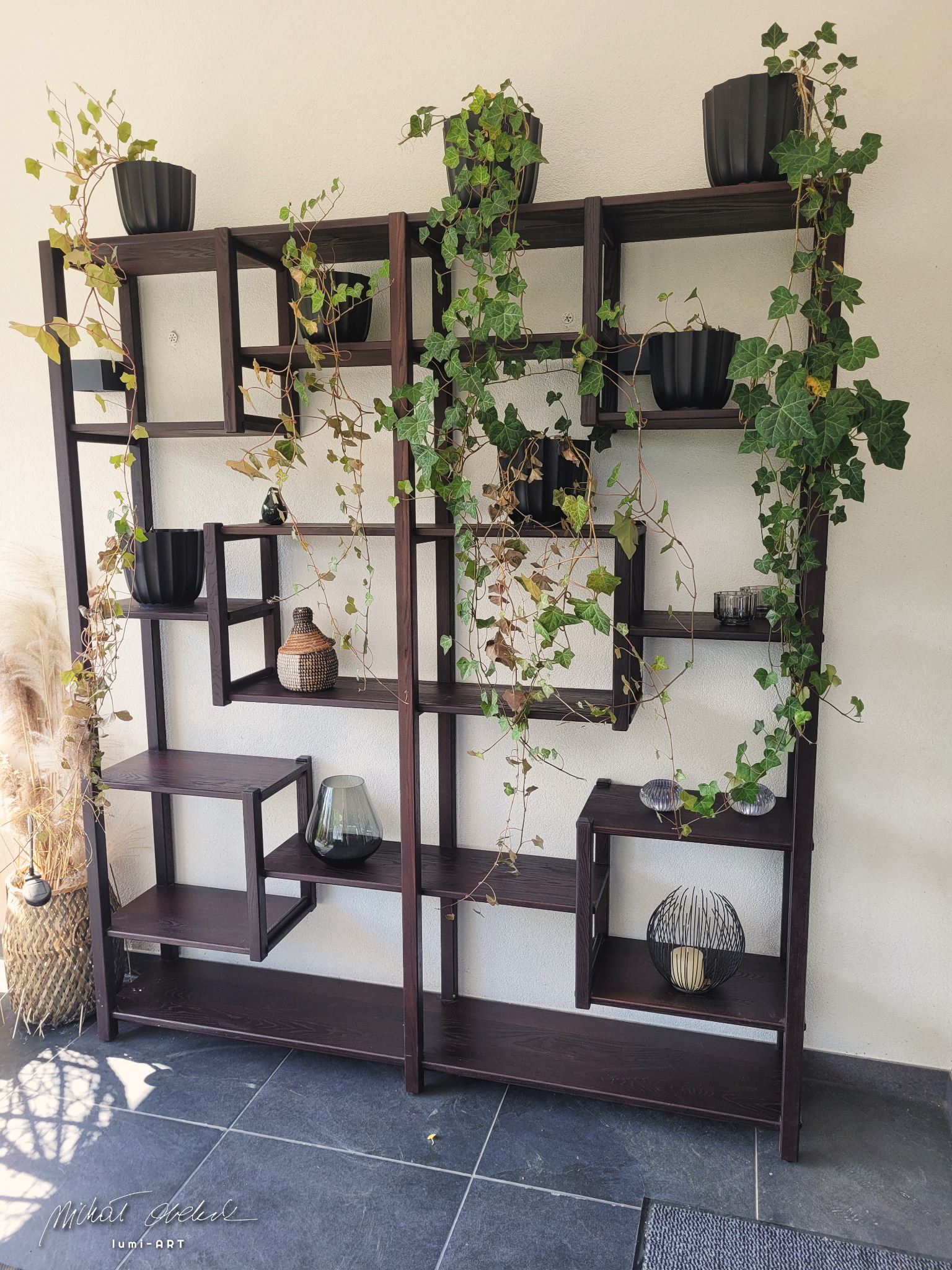 Ręcznie robiony regał z ciemnego drewna, asymetryczny układ półek, dekoracje roślinne w doniczkach i ozdobne przedmioty, nowoczesny design.
