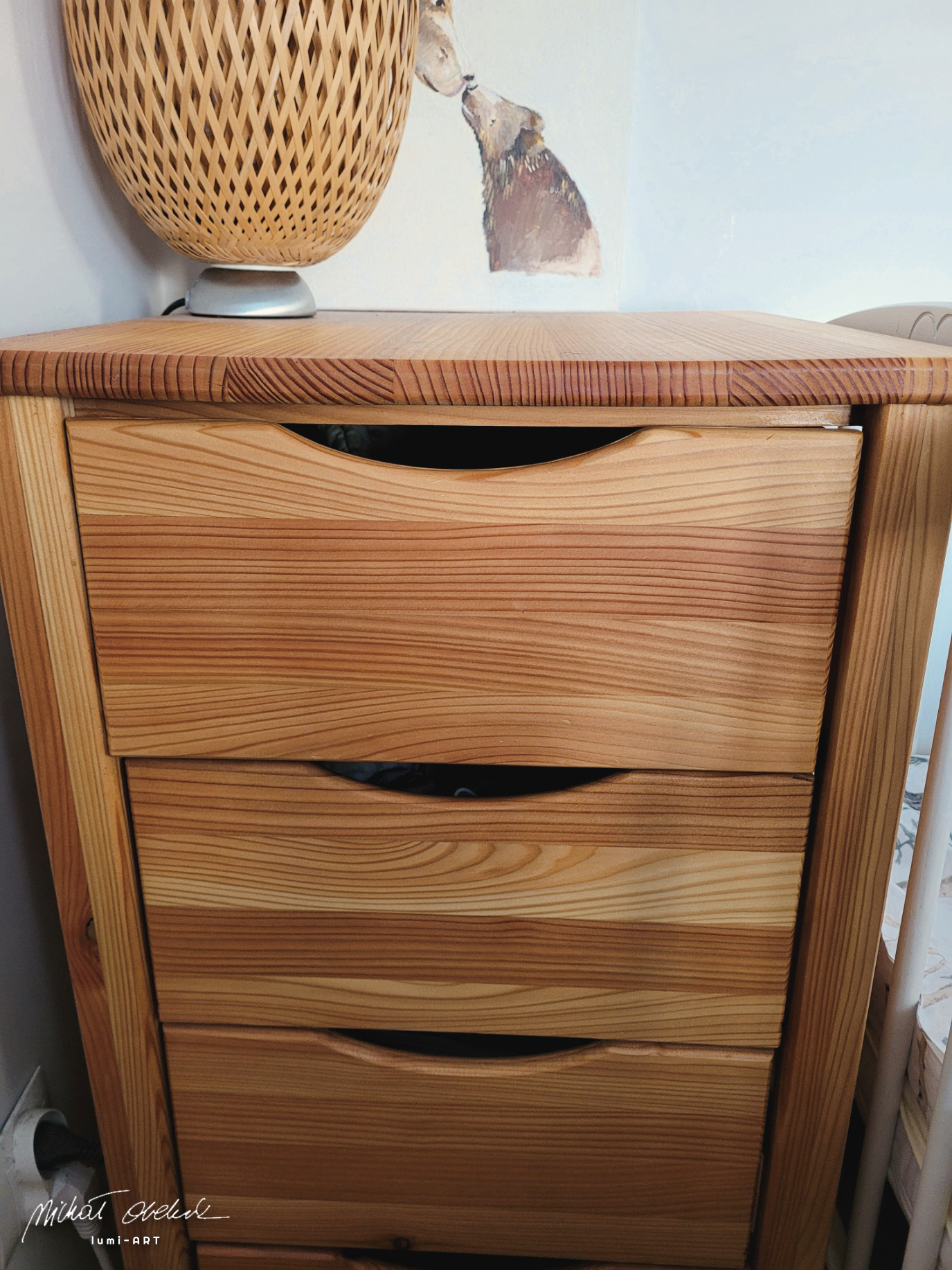 Drewniana komoda z trzema szufladami o wyraźnym usłojeniu, z lampką nocną i obrazkiem z motywem zwierzęcym w tle, ukazana w zbliżeniu.