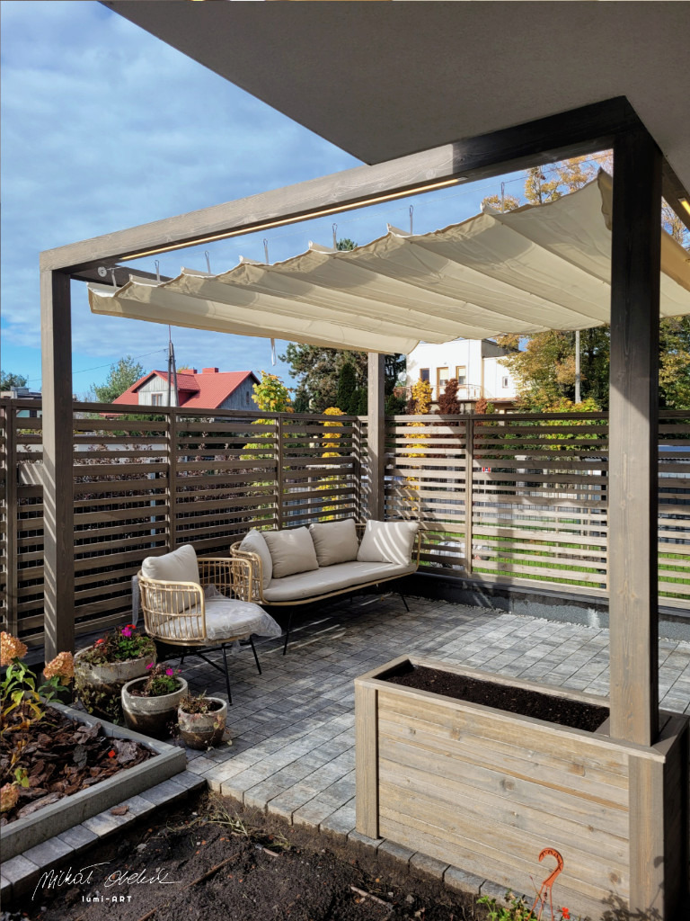 Nowoczesna pergola z beżową, zwijaną markizą, otoczona drewnianym ogrodzeniem i roślinami w donicach. Wygodny zestaw wypoczynkowy na szarym bruku.