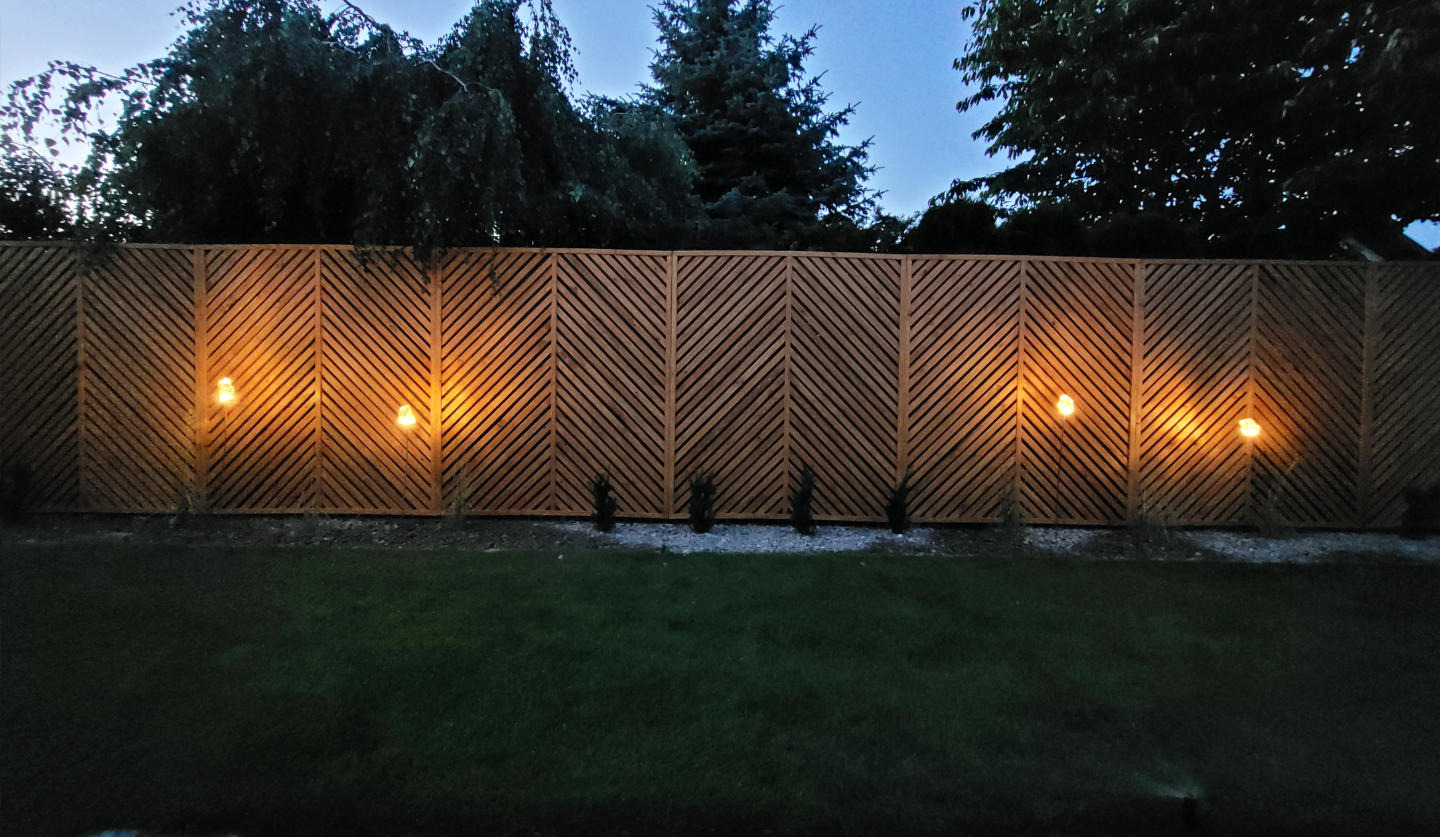 Drewniane ogrodzenie z ukośnymi listwami, oświetlone lampami ogrodowymi, tworzące ciepłą atmosferę w ogrodzie o zmierzchu. Widoczne drzewa w tle.