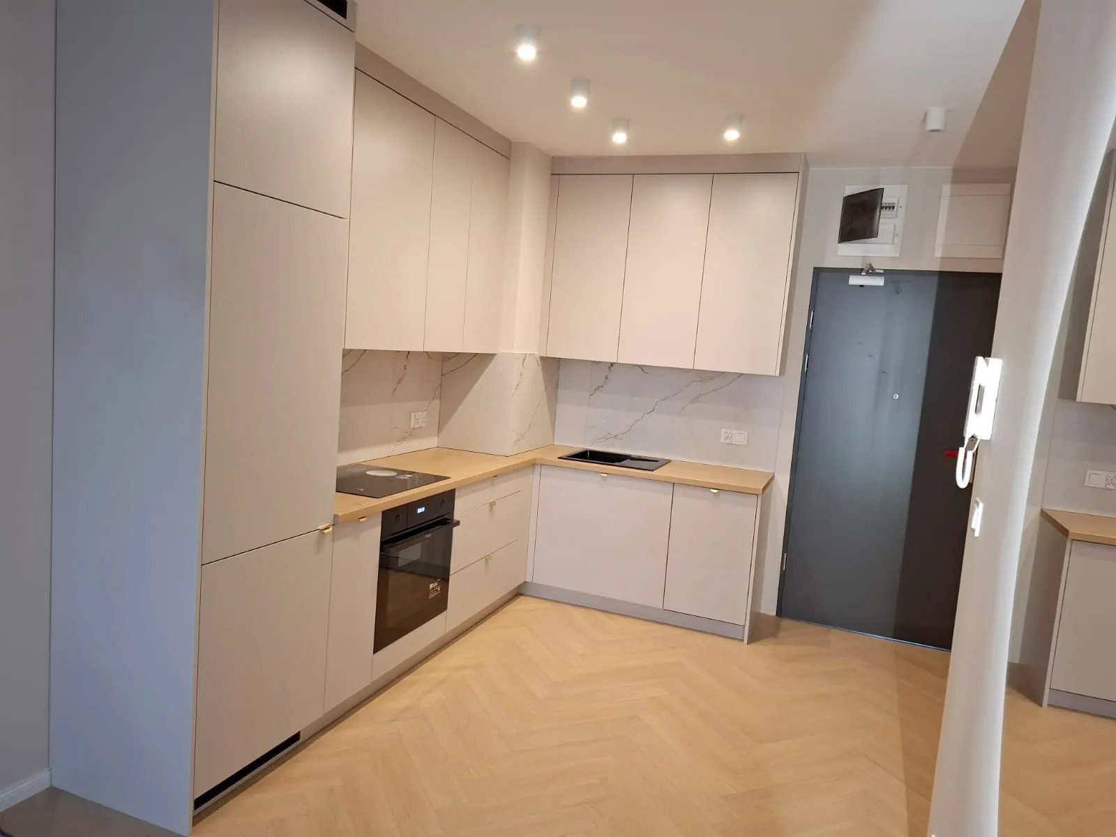 Jasna kuchnia z drewnianym blatem i marmurowym backsplashiem. Minimalistyczne szafki bez uchwytów, płyta indukcyjna i piekarnik. Parkiet w jodełkę na podłodze.