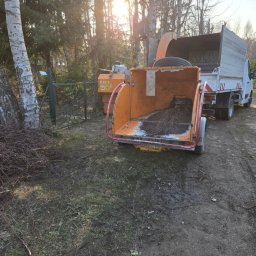 M-LIFT - Pomarańczowy rębak do drewna z otwartą komorą, gotowy do pracy, obok stosu świeżo ściętych gałęzi. W tle drzewa i słońce, z tyłu biała ciężarówka.