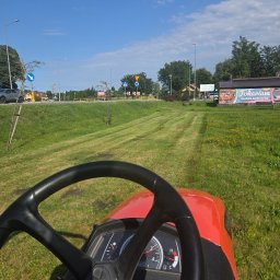 M-LIFT - Widok zza kierownicy czerwonej kosiarki samojezdnej, koszącej trawę w równe pasy. W tle droga, znaki i budynek 'Fokarium Sarbsk koło Łeby'.