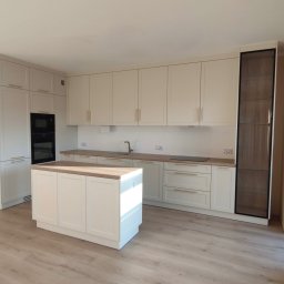 Meblorak Meble na wymiar - Jasna kuchnia z wyspą i drewnianym blatem. Fronty szafek w stylu klasycznym, złote uchwyty. W tle zabudowa piekarnika i wysoka szafa z przeszkleniami.