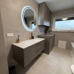 Meblorak Meble na wymiar - Nowoczesna łazienka z szarą szafką pod umywalką, okrągłym lustrem z podświetleniem LED i wiszącą szafką. Minimalistyczny design, neutralna kolorystyka.