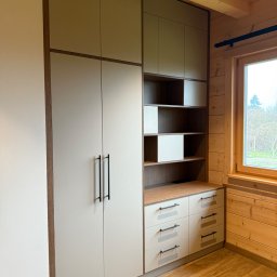 Meblorak Meble na wymiar - Jasna szafa wnękowa z szufladami i półkami, czarne uchwyty, drewniane wykończenie, wnętrze domu z drewnianymi ścianami i podłogą, widok na okno.