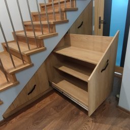 Meblorak Meble na wymiar - Drewniane schody z zabudową meblową pod skosami, szuflady z półkami na metalowych prowadnicach. Widoczna drewniana podłoga i fragment drzwi.