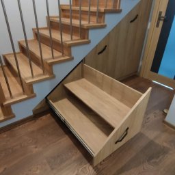 Meblorak Meble na wymiar - Drewniane schody z zabudową meblową pod skosami, wysuwana szuflada z dwoma półkami. Metalowe barierki, drewniana podłoga. Funkcjonalne wykorzystanie przestrzeni.