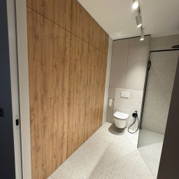 Meblorak Meble na wymiar - Nowoczesna łazienka z drewnianą szafą na wymiar, białą toaletą i szarą kabiną prysznicową. Minimalistyczny design, jasne oświetlenie punktowe.