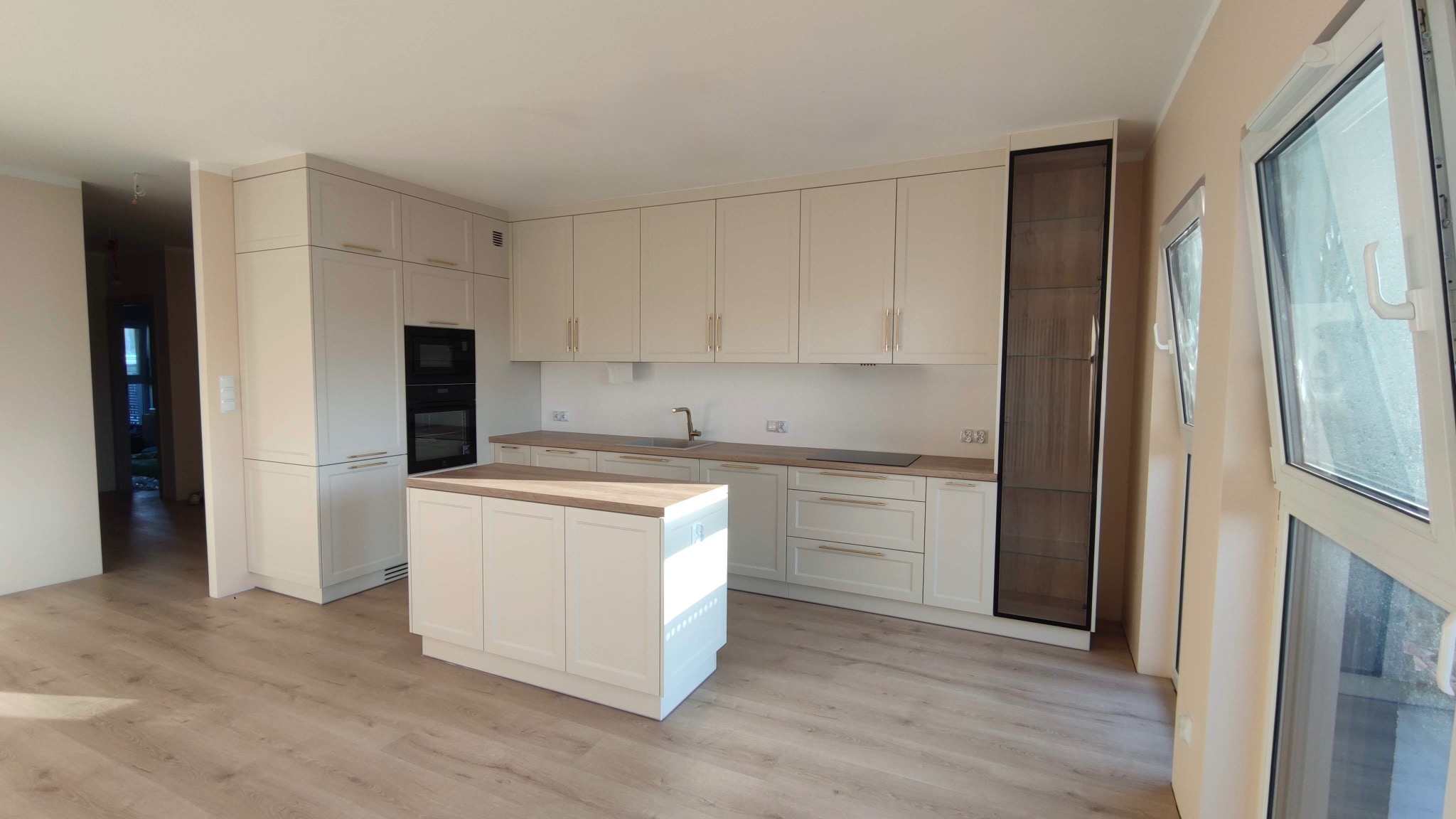 Jasna kuchnia z wyspą i drewnianym blatem. Fronty szafek w stylu klasycznym, złote uchwyty. W tle zabudowa piekarnika i wysoka szafa z przeszkleniami.